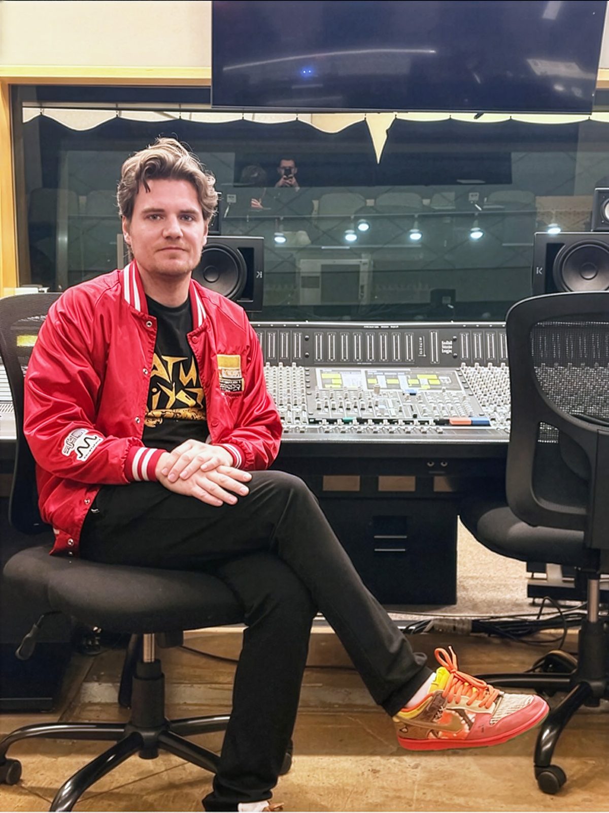 Joshua M. Lutz in a red varsity jacket seated at a mixing console in a recording studio control room