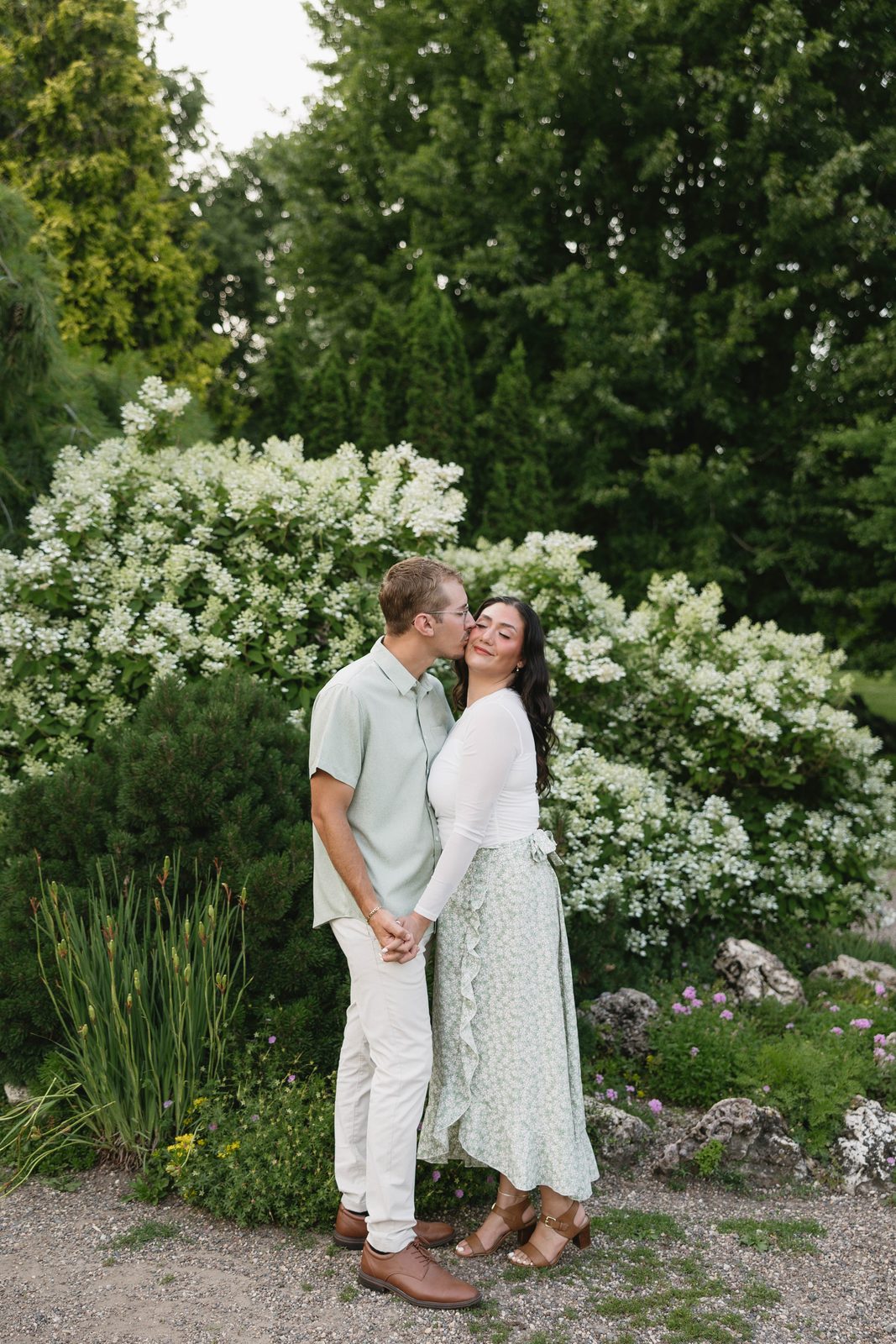 A couple holding hands in front of white hydrangeas, the groom kissing the bride on the cheek, photographed by Captured by Kaitlyn
