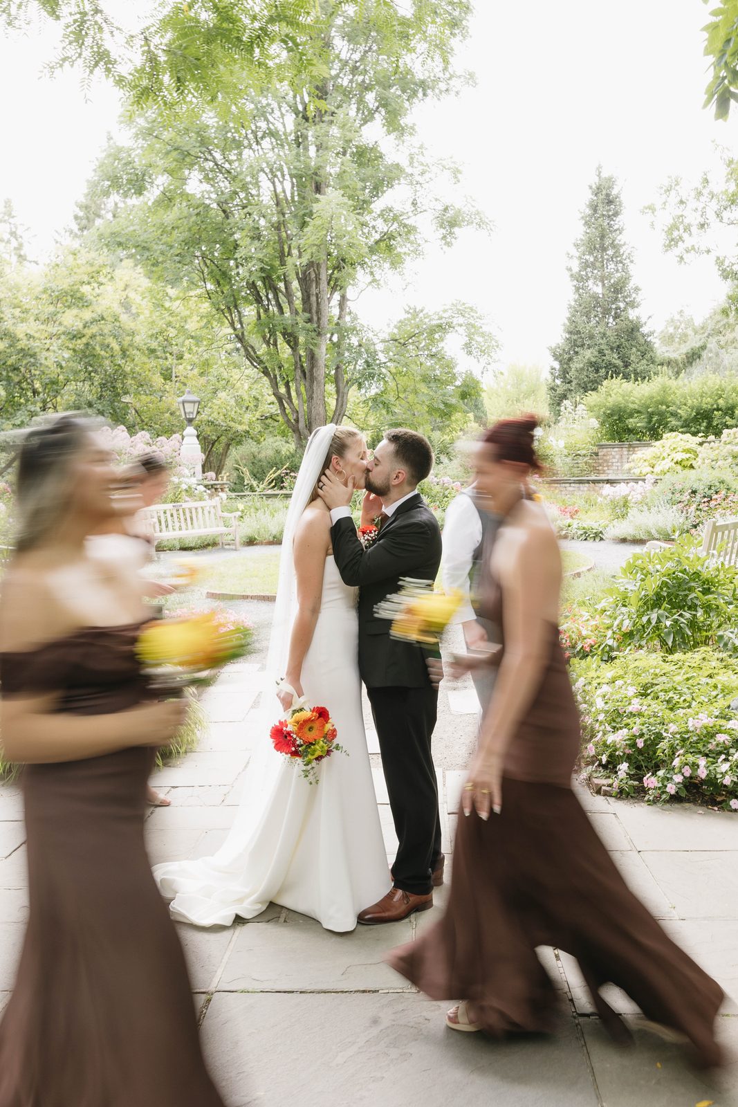 A bride and groom kissing in a garden while bridesmaids in brown dresses walk past in motion blur, photographed by Captured by Kaitlyn