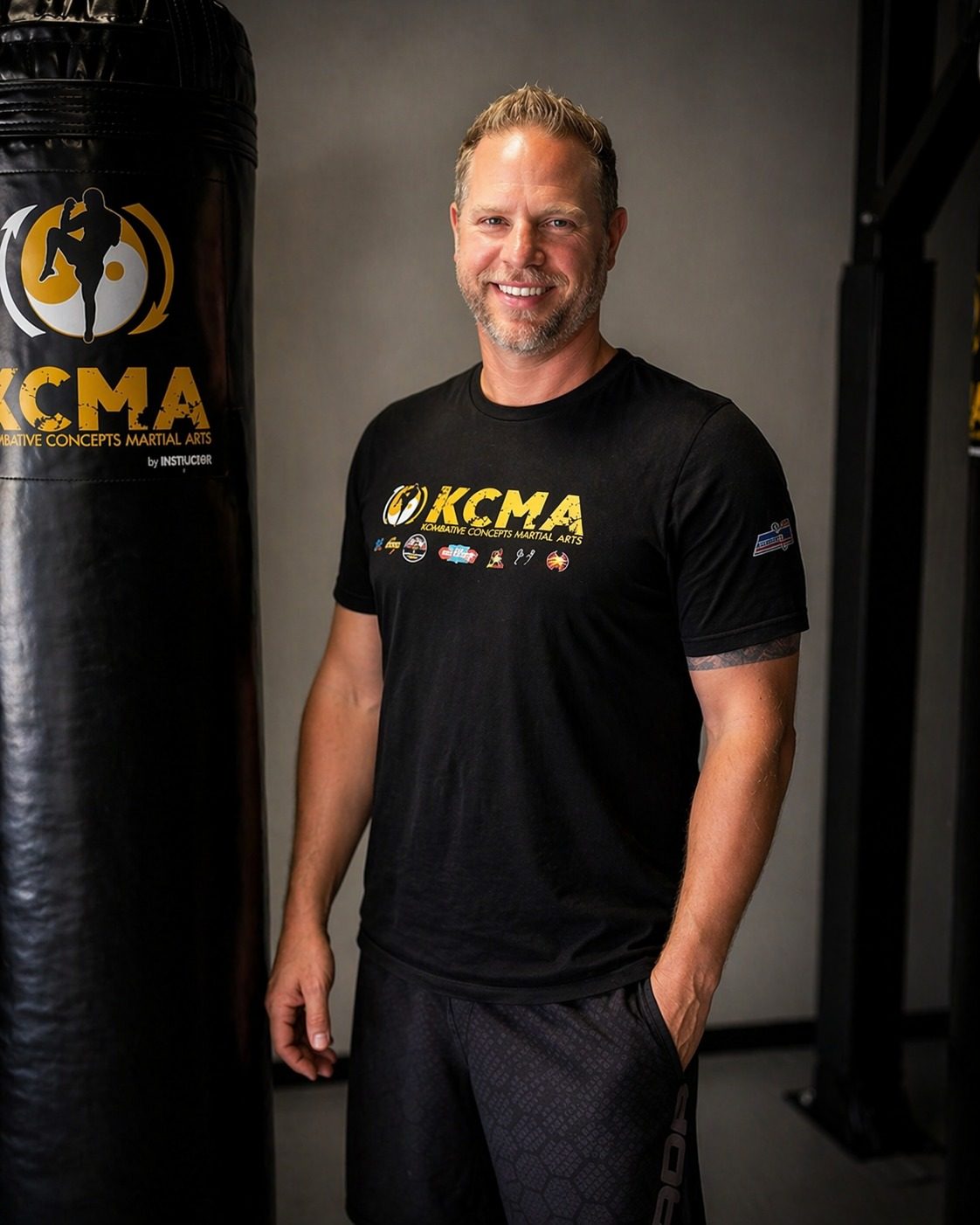 Keith Kirkpatrick, founder of Kombative Concepts Martial Arts, smiling beside a heavy bag in a KCMA T-shirt