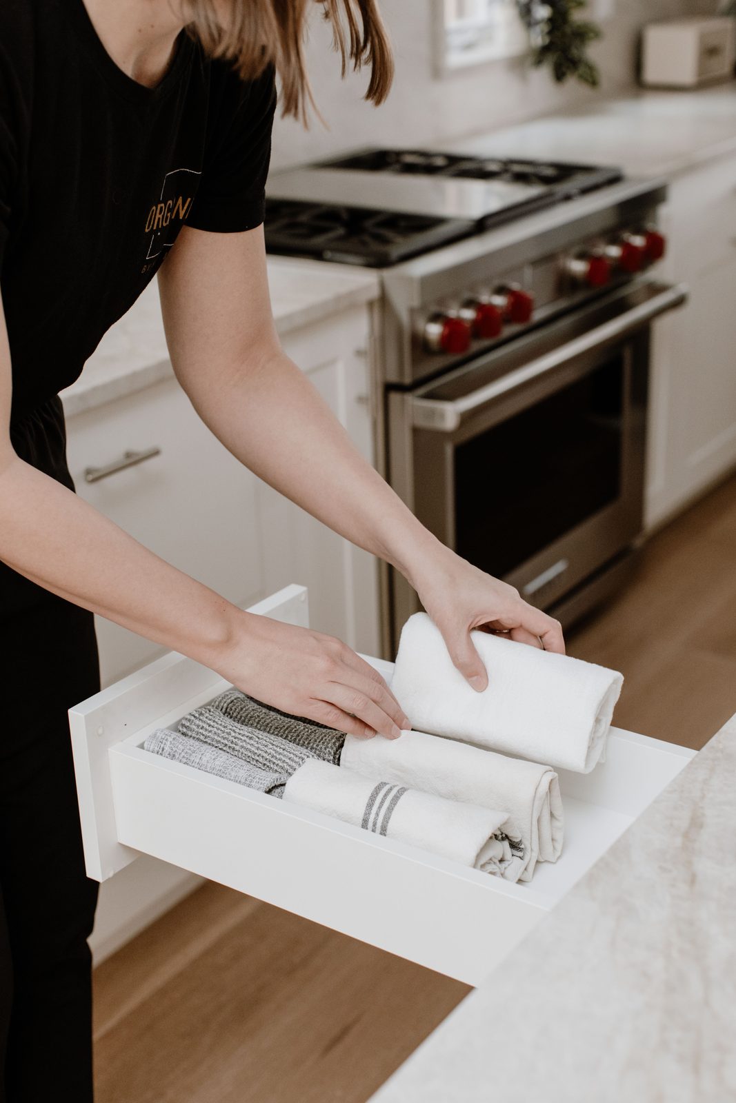 An Organized by Keli & Co. organizer rolling kitchen towels into a white drawer beside a stainless steel range