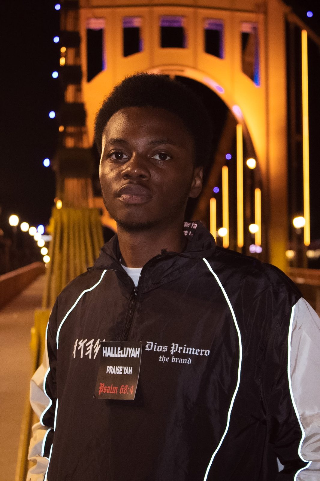 Keysean Jenkins, founder of Dios Primero, in a black Dios Primero jacket reading Halleluyah Praise Yah on a lit bridge in Pittsburgh at night