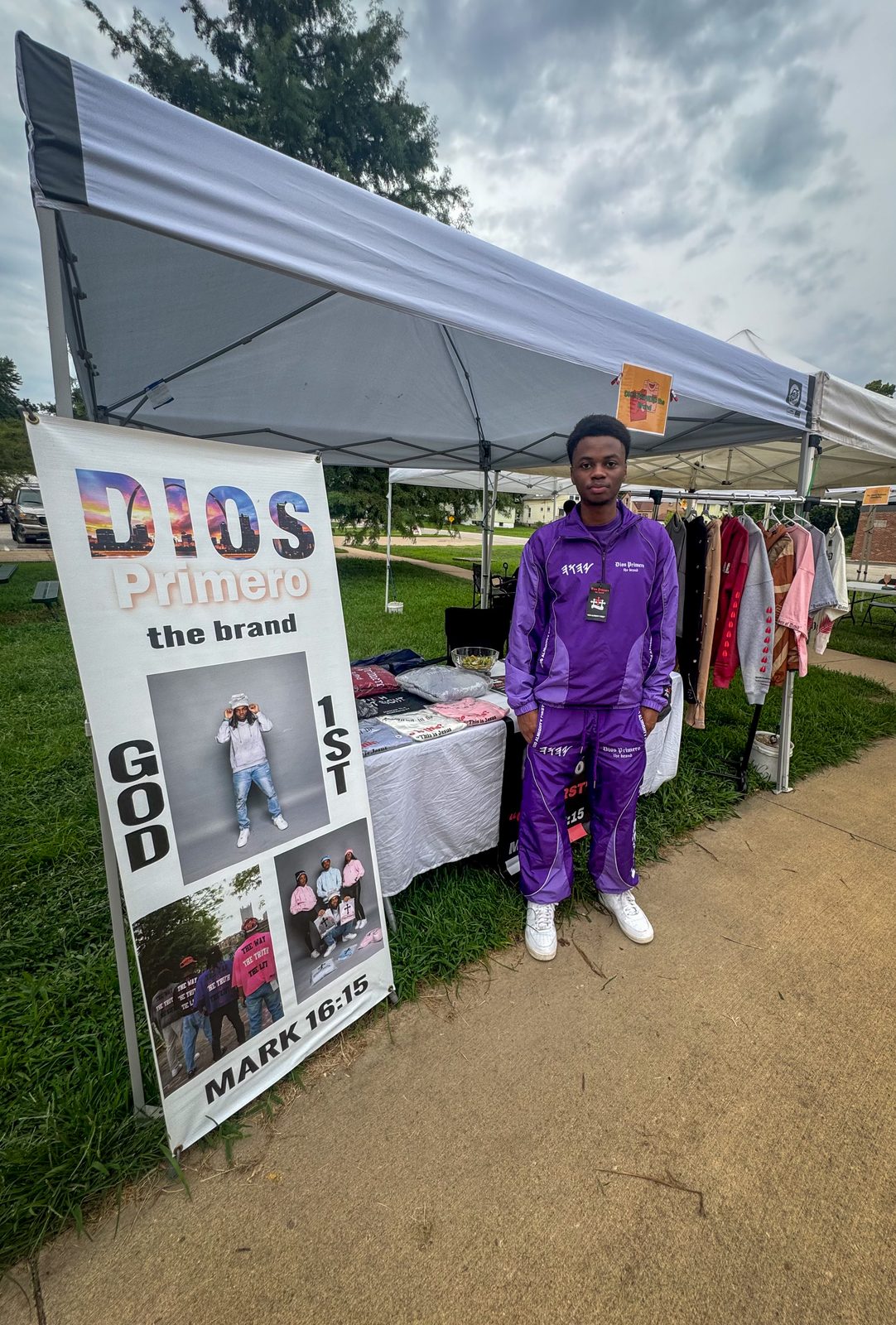 Keysean Jenkins in a purple Dios Primero tracksuit at the brand's market tent, beside a Dios Primero banner and a rack of clothing