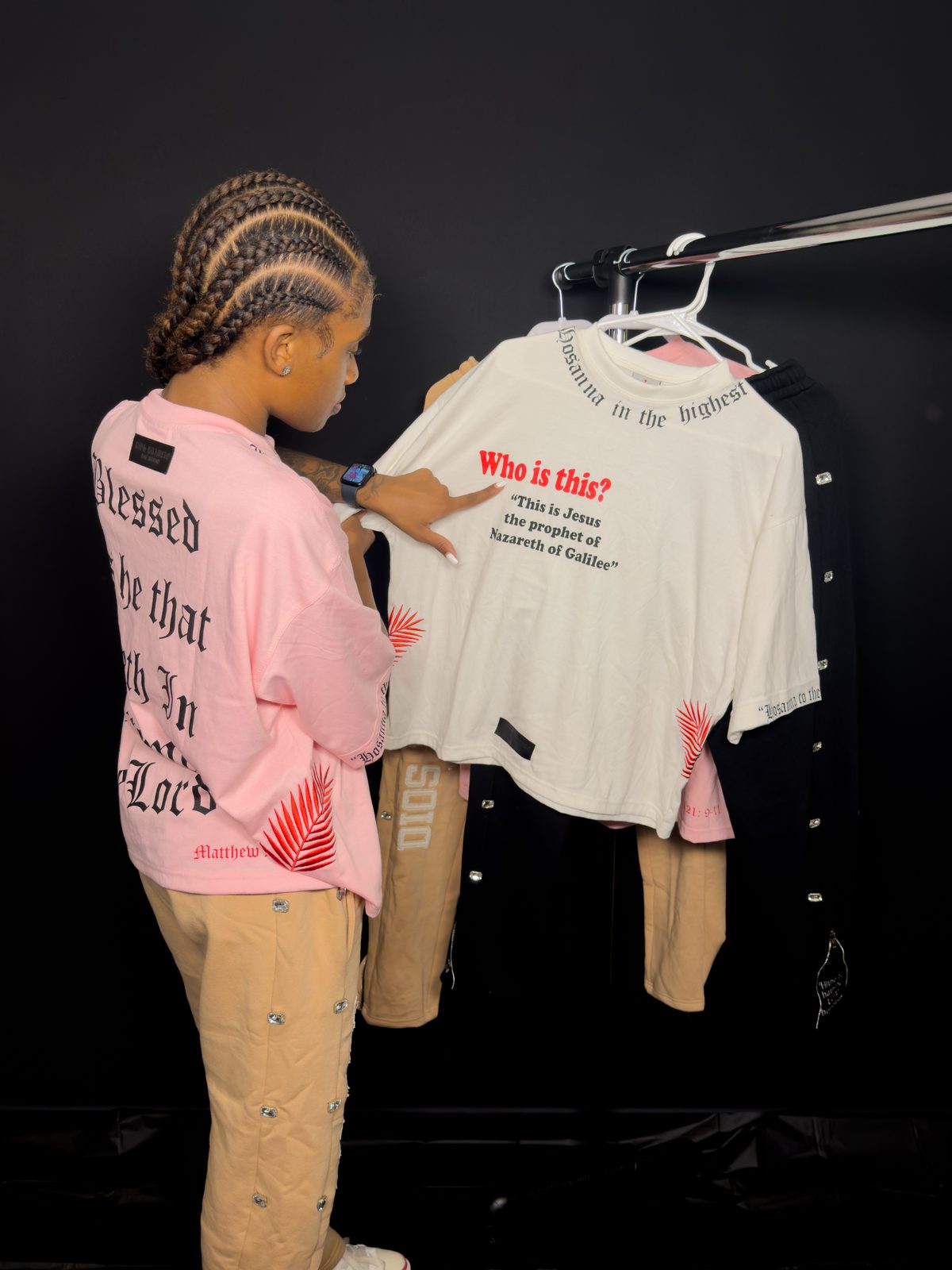 A shopper in a pink Dios Primero tee browsing a rack of the brand's shirts, holding a white tee printed with Who is this? This is Jesus the prophet of Nazareth of Galilee