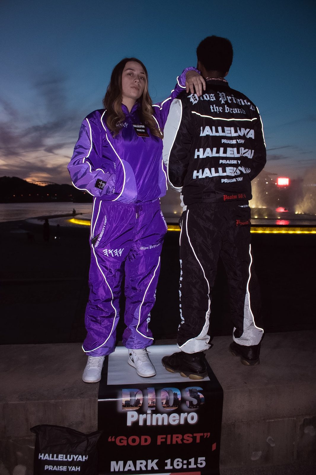 Two models in reflective purple and black Dios Primero tracksuits standing on a podium at dusk above a banner reading Dios Primero, God First, Mark 16:15