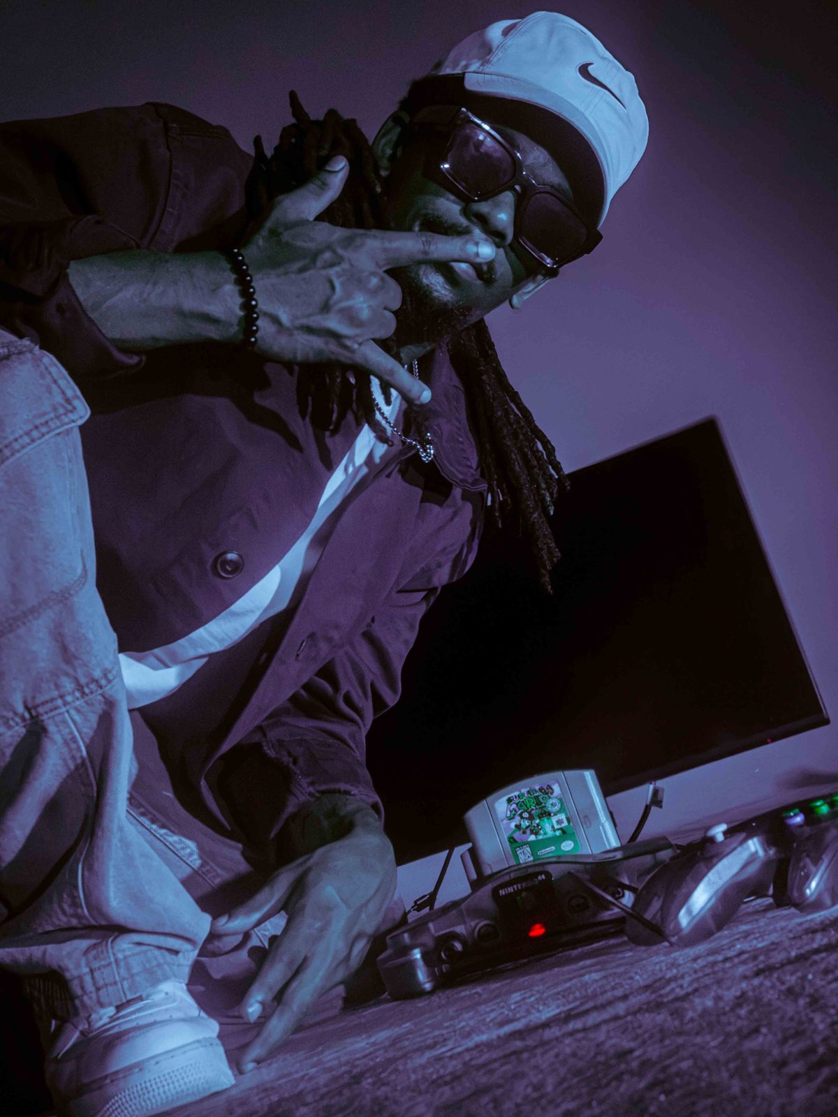 Khalid Thornton throwing up a hand sign in a purple-lit studio, with a Nintendo 64 behind him