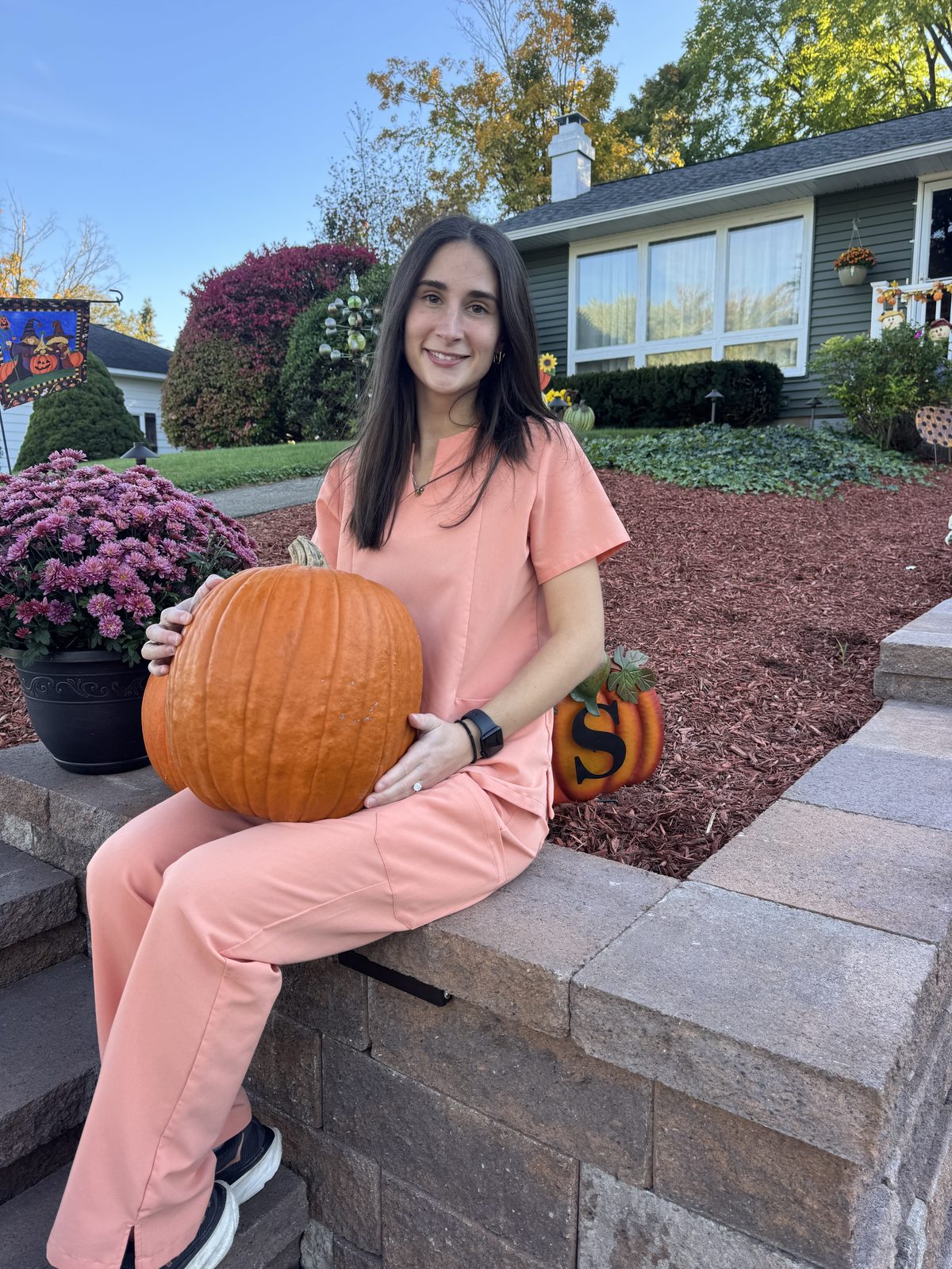 Kristen Sellars in peach scrubs sitting on a stone wall holding a large pumpkin, with fall mums beside her