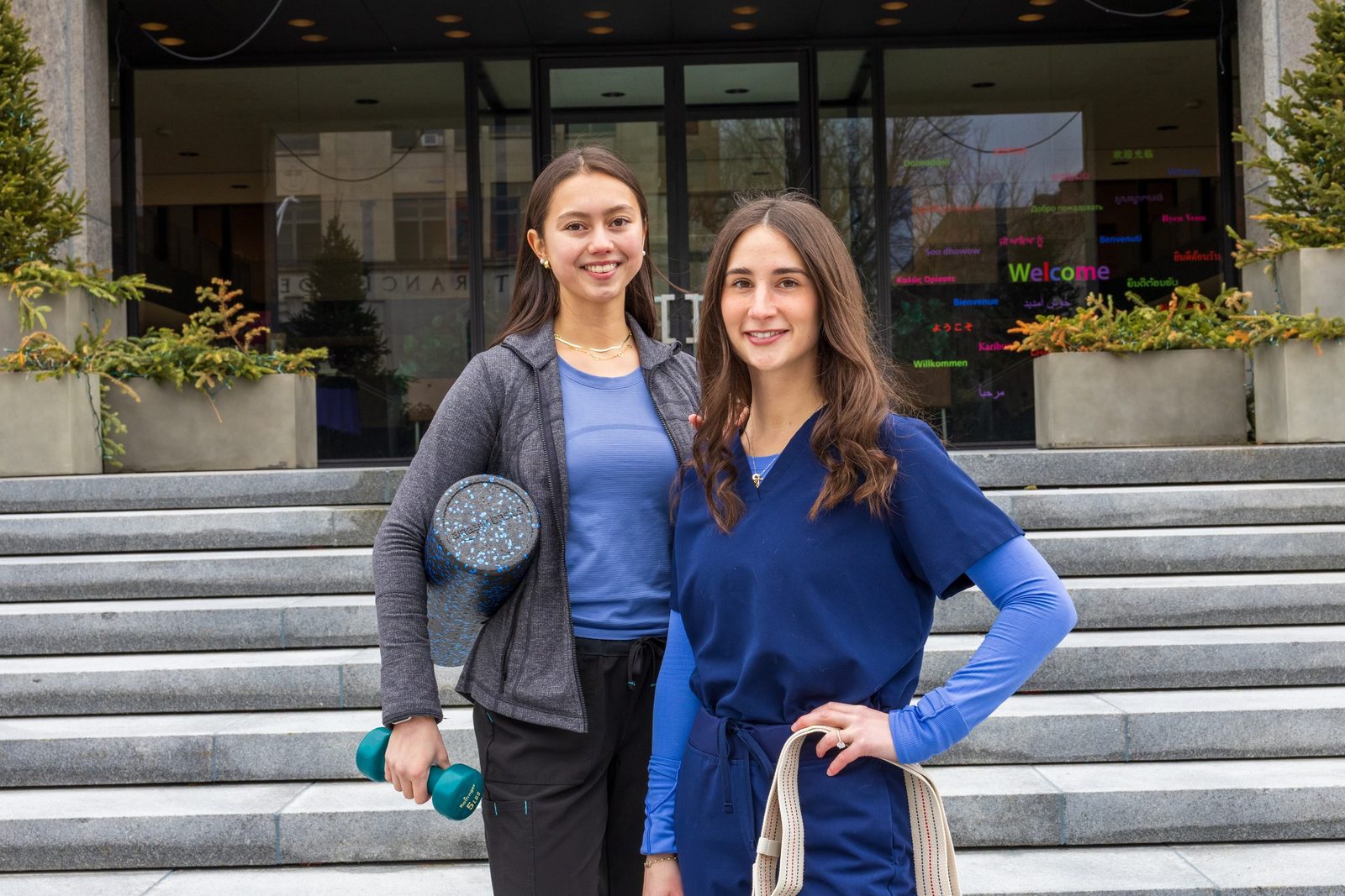 Kristen Sellars in blue scrubs with her OT Insider Podcast co-host Kiana, a physical therapist, on the steps outside a gym