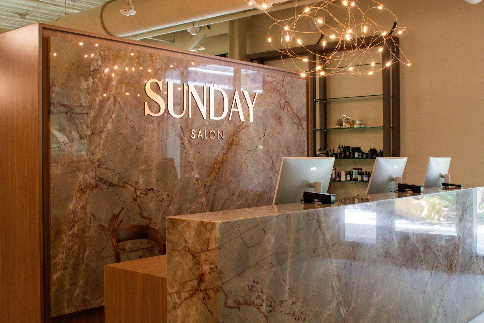 The marble reception desk at Sunday Salon with the salon's name in brass lettering beneath a sculptural string-light chandelier