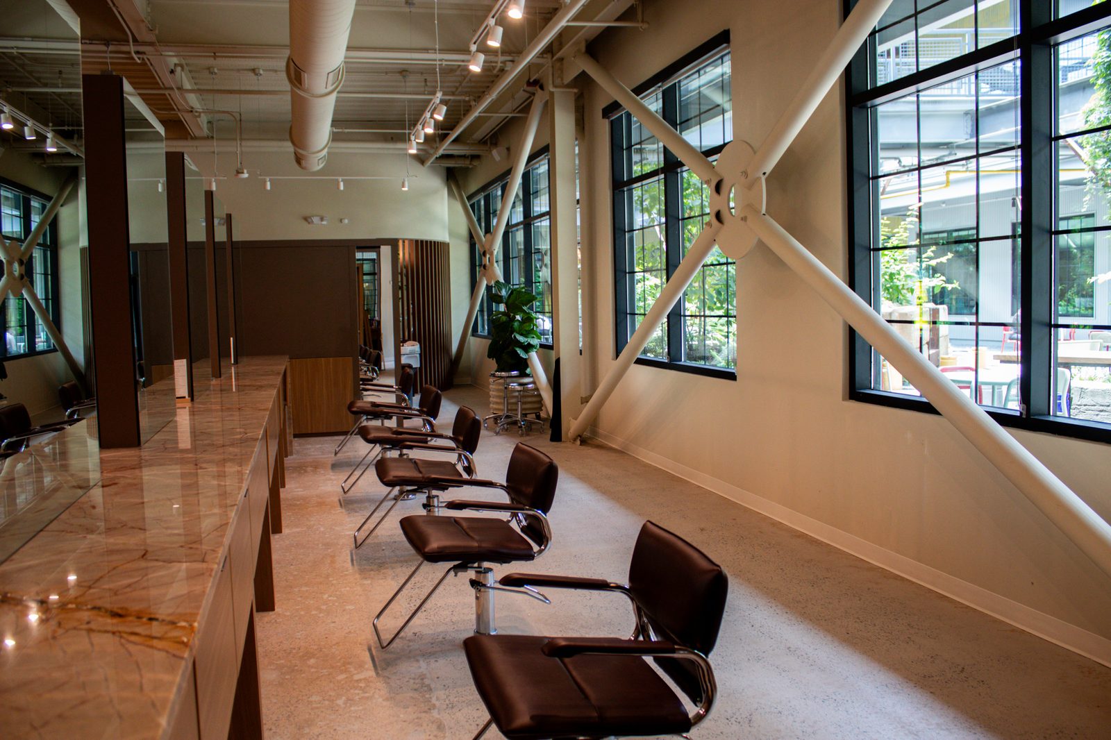 A row of styling chairs along marble counters beneath tall black-framed factory windows at Sunday Salon