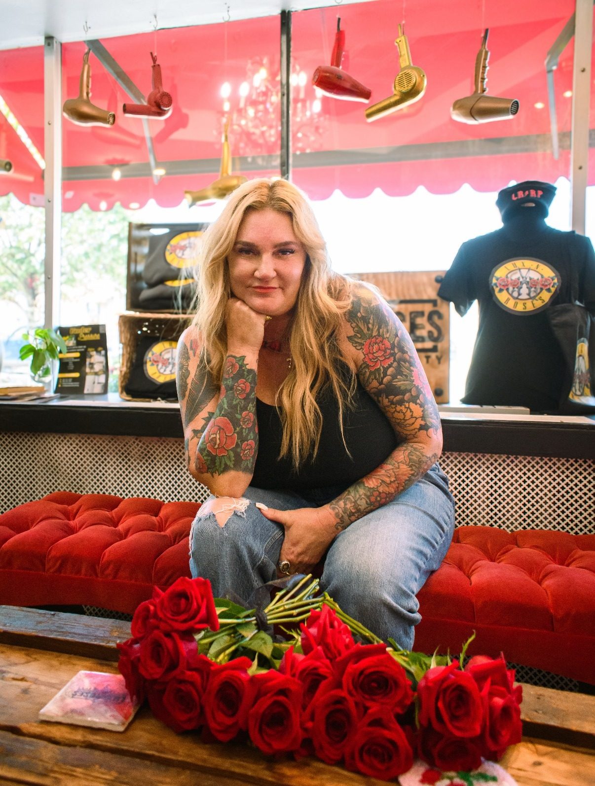 Machele Alessandrine, owner of Lux and Roses, sitting in her salon window with a bouquet of red roses, hair dryers hanging overhead