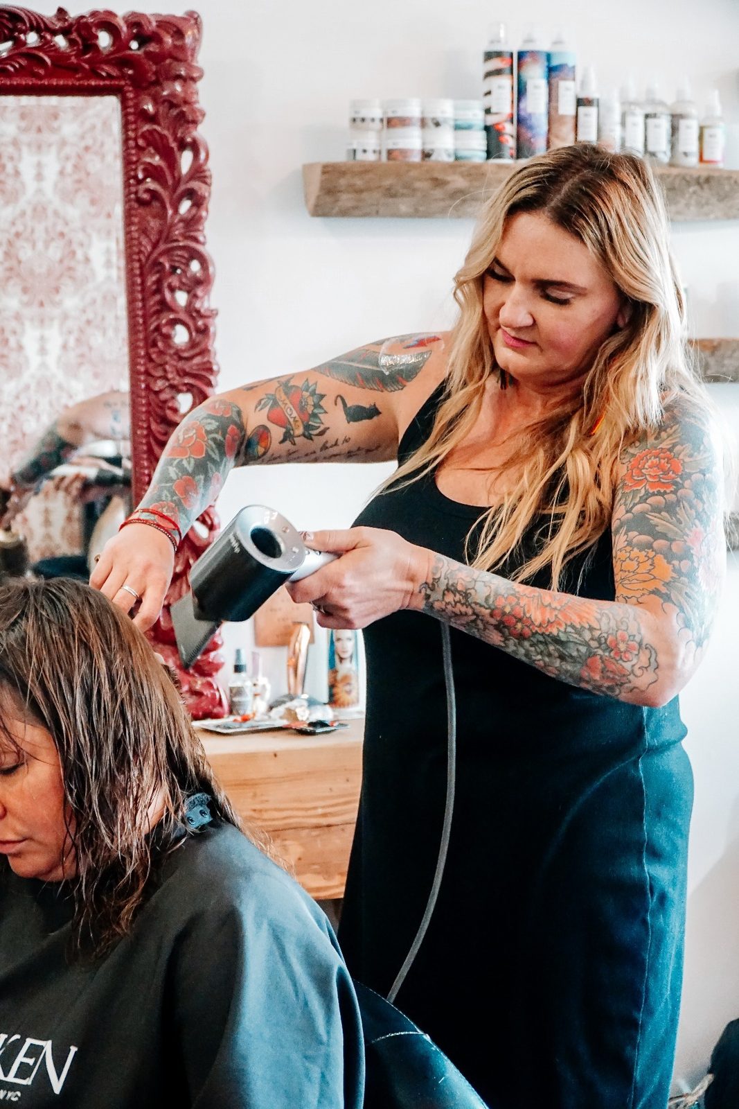 Machele Alessandrine blow-drying a client's hair at a station with an ornate red mirror