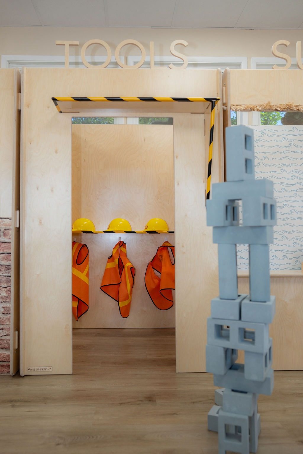A construction pretend-play area at Coastal Kids Playspace with yellow hard hats and orange safety vests on hooks beside stacked foam building blocks