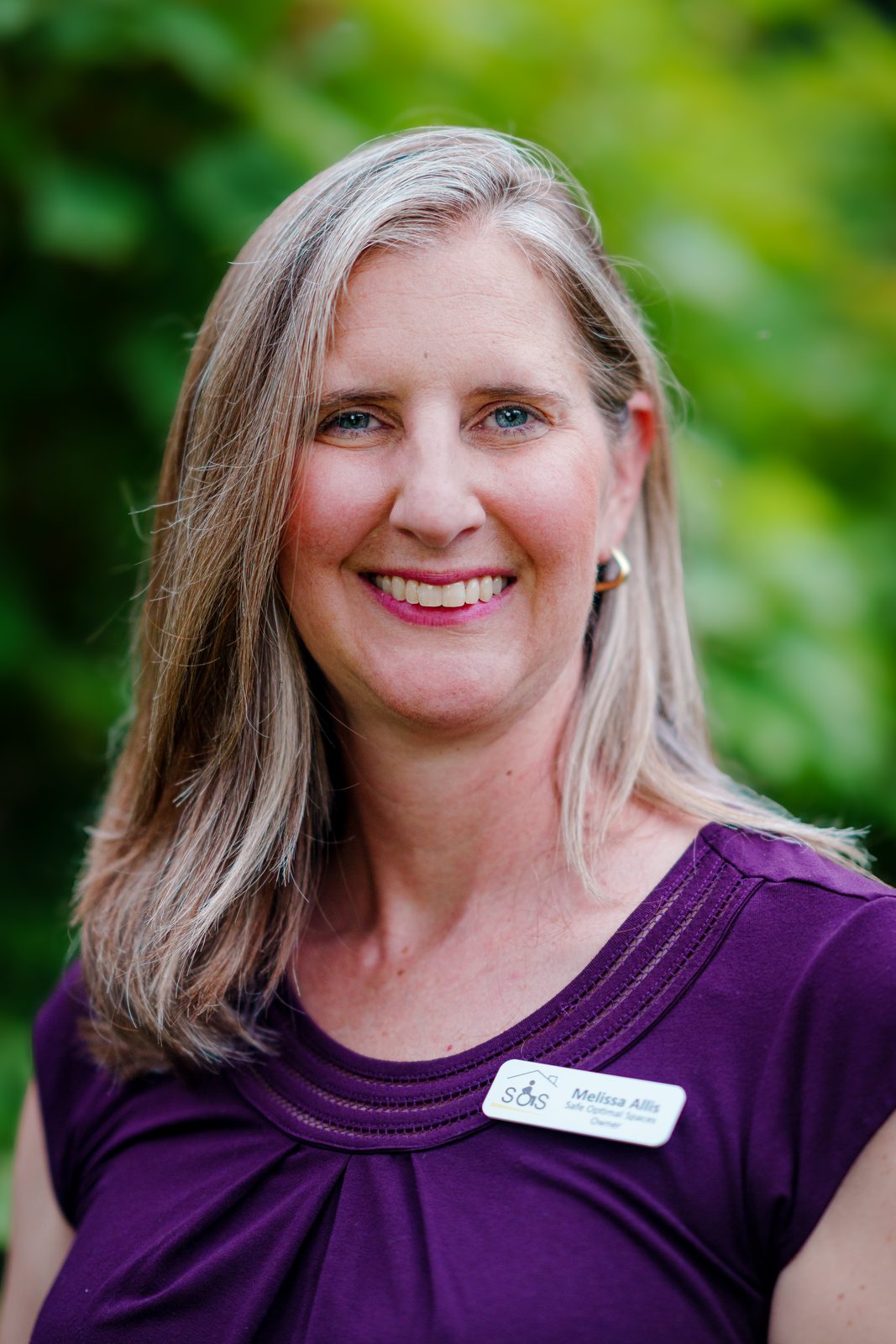 Melissa Allis, founder of Safe Optimal Spaces, smiling in a purple top with a name badge against green foliage