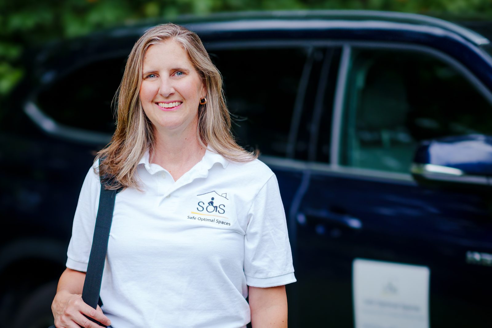 Melissa Allis in a white Safe Optimal Spaces polo with a bag over her shoulder, standing beside her car