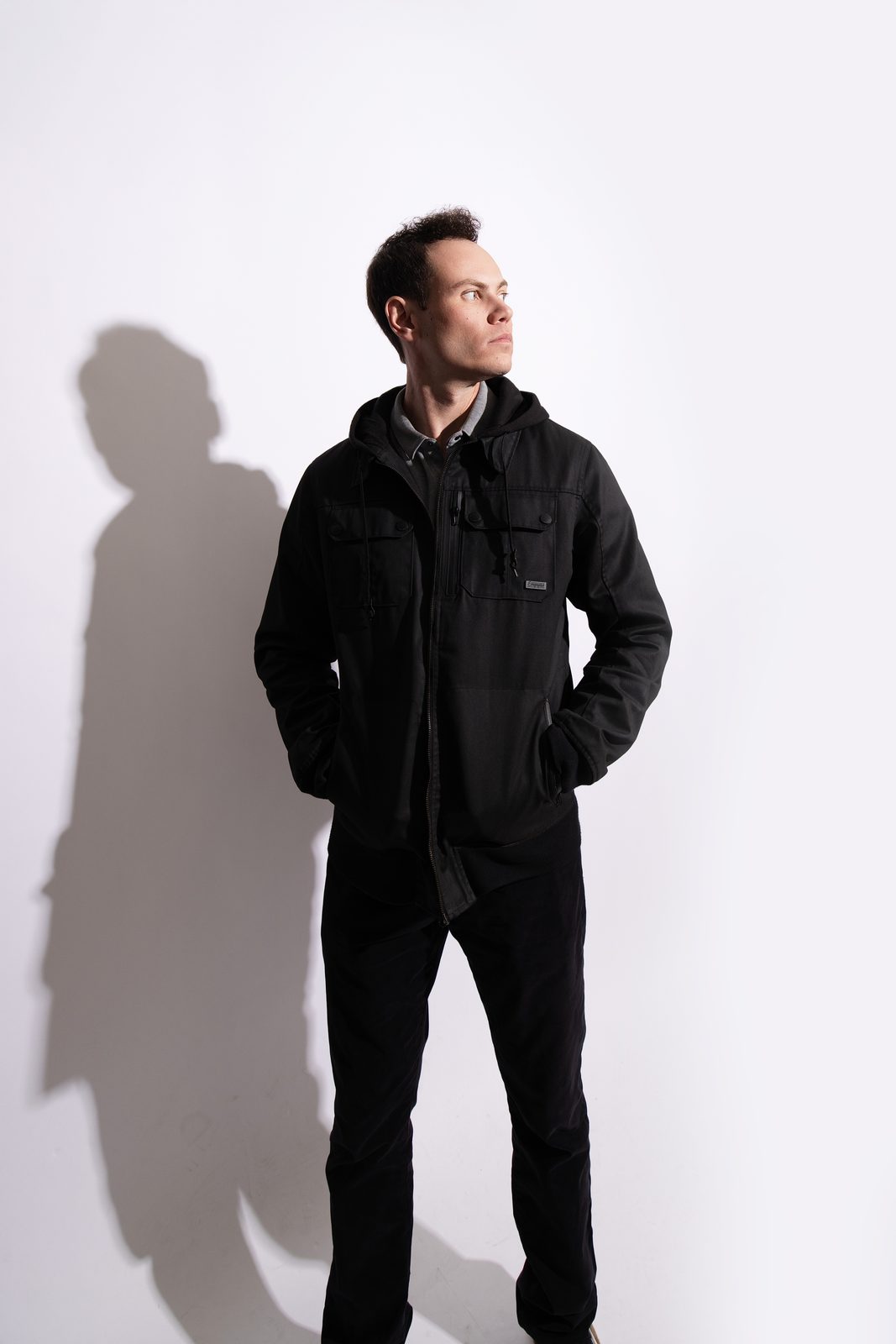 Michael Cecchinelli standing in a black hooded jacket and black jeans, looking off to the side against a white studio wall