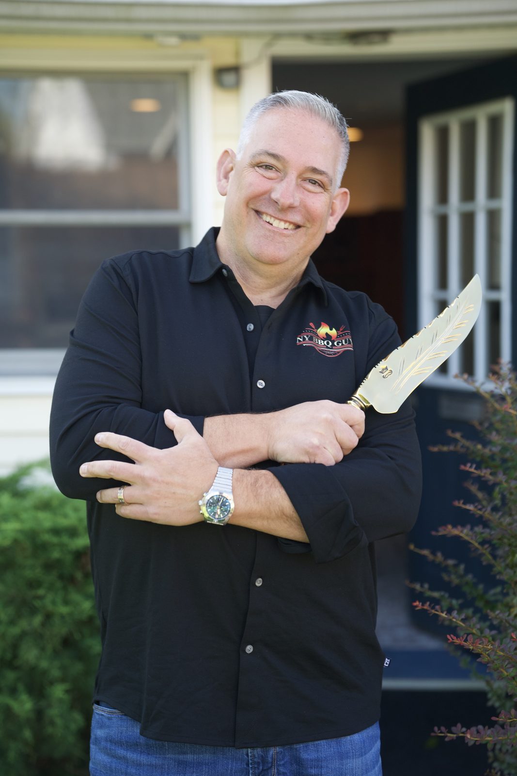 Mike Riddle, founder of Own The Fire, smiling with arms crossed and holding an engraved chef's knife