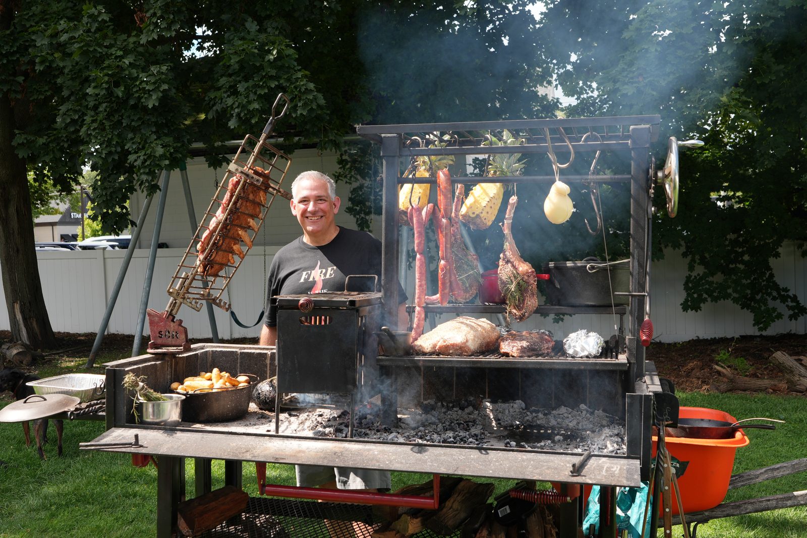 Mike Riddle smiling behind a hand-built Santa Maria grill loaded with meats and vegetables over live coals
