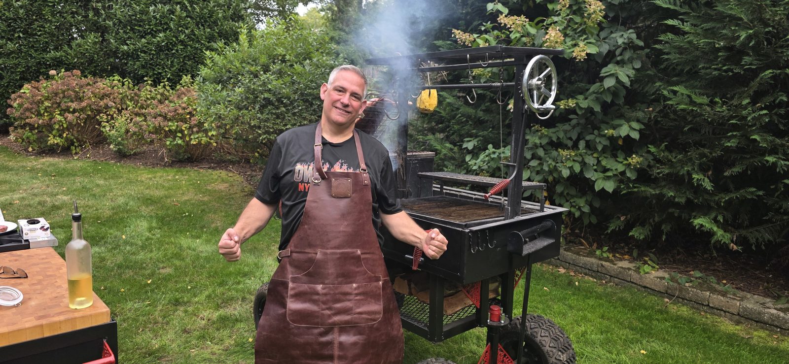 Mike Riddle in a leather apron in front of a smoking custom-fabricated grill