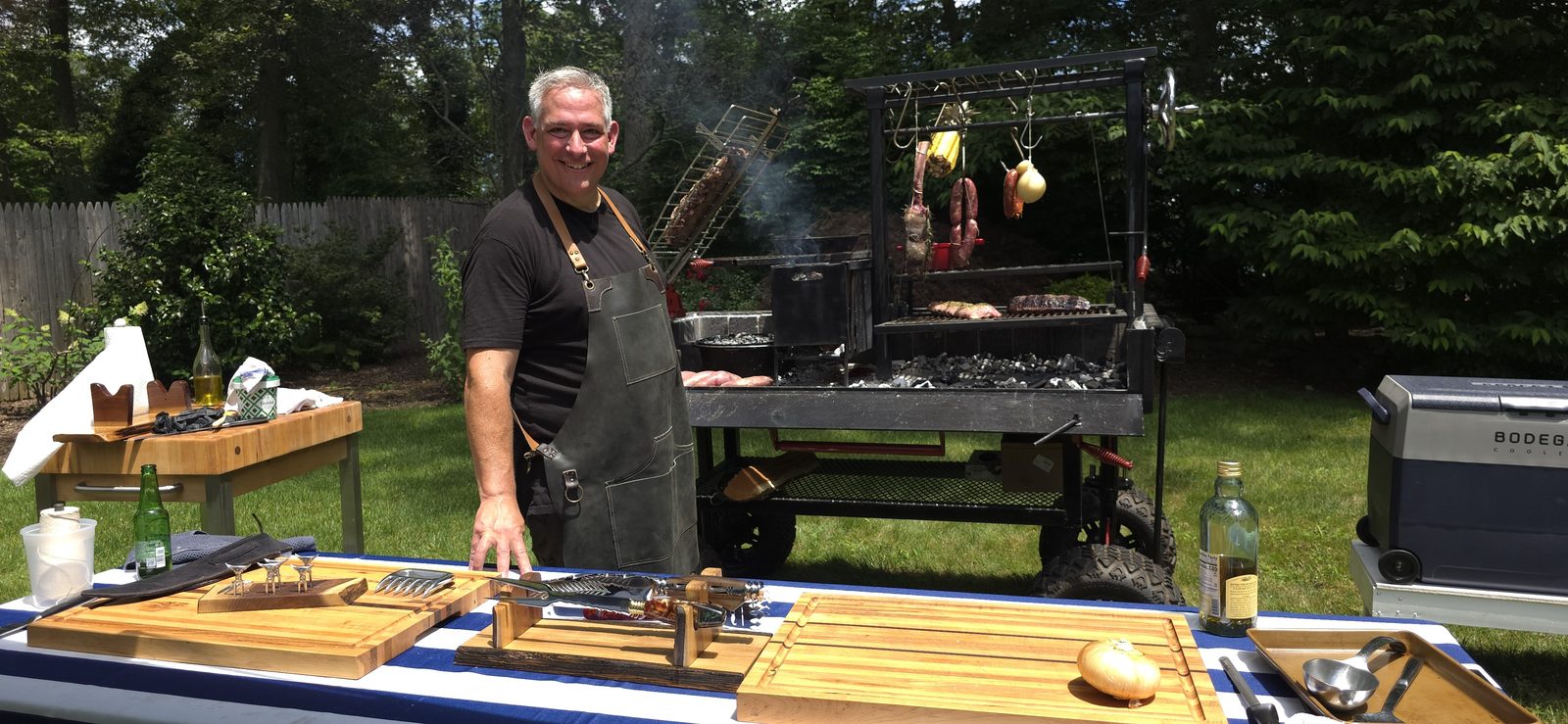 Mike Riddle at an outdoor prep station with cutting boards and knives, a custom grill cooking behind him