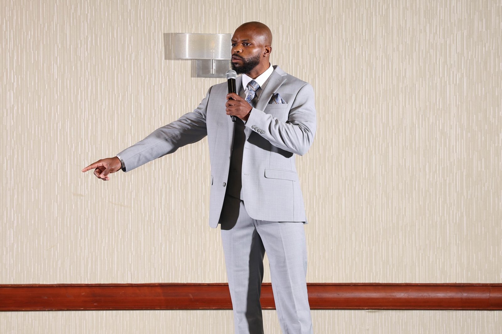 Milton Putman speaking on stage with a microphone in a grey suit, pointing to the audience