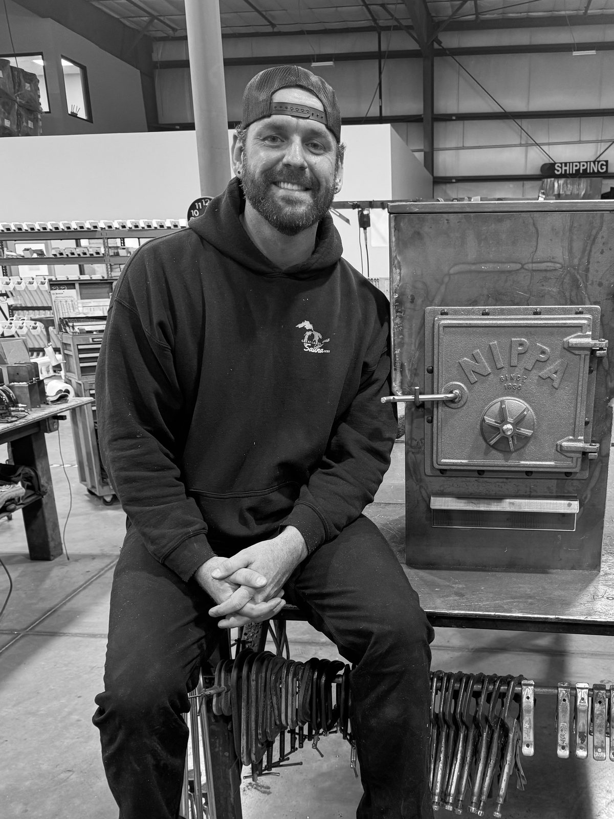 Philip Casteleyn, owner of Nippa Sauna Stoves, smiling in a black hoodie and backwards cap beside a Nippa wood-burning stove
