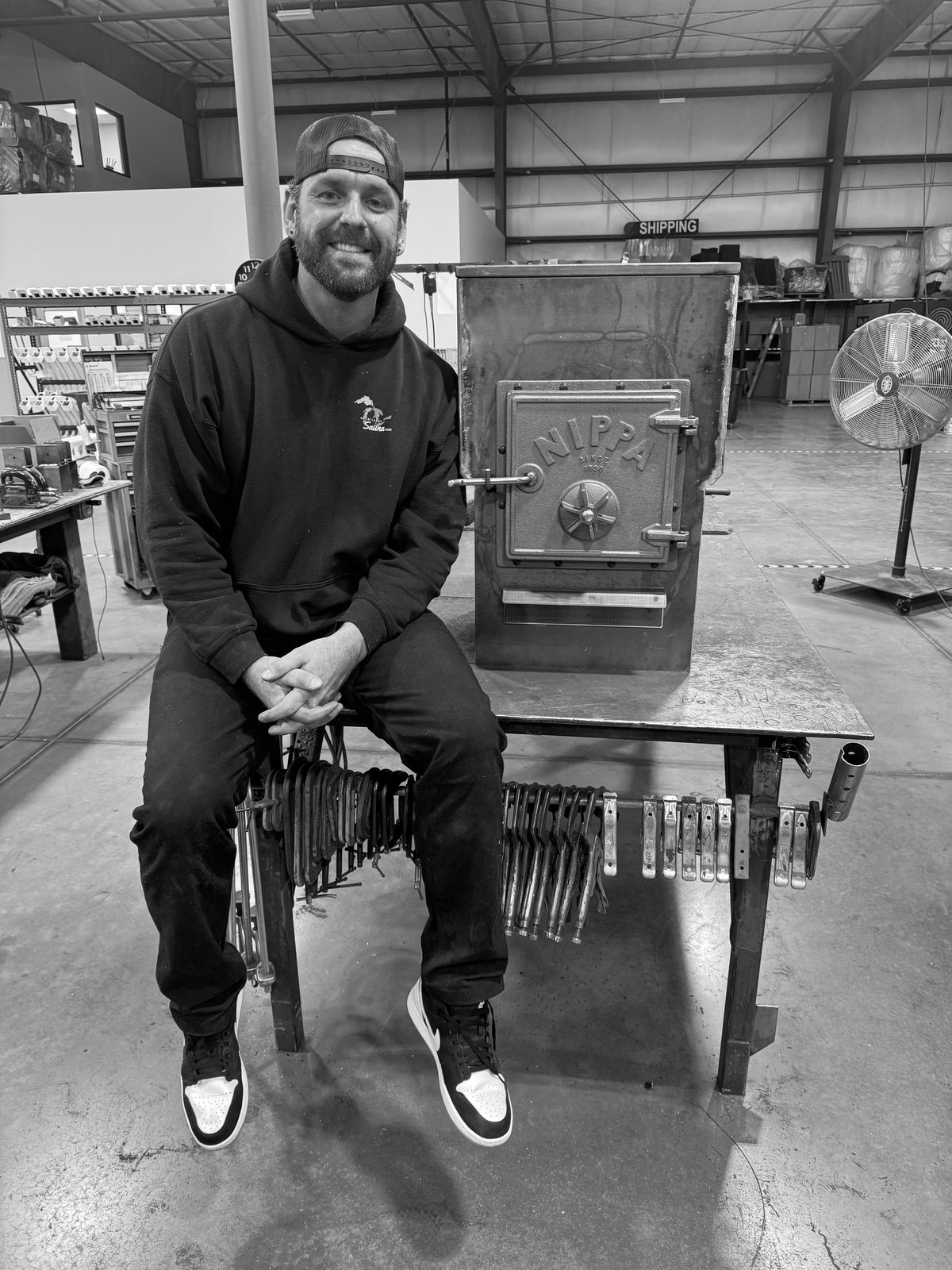 Philip Casteleyn seated on a steel workbench next to a Nippa sauna stove on the factory floor