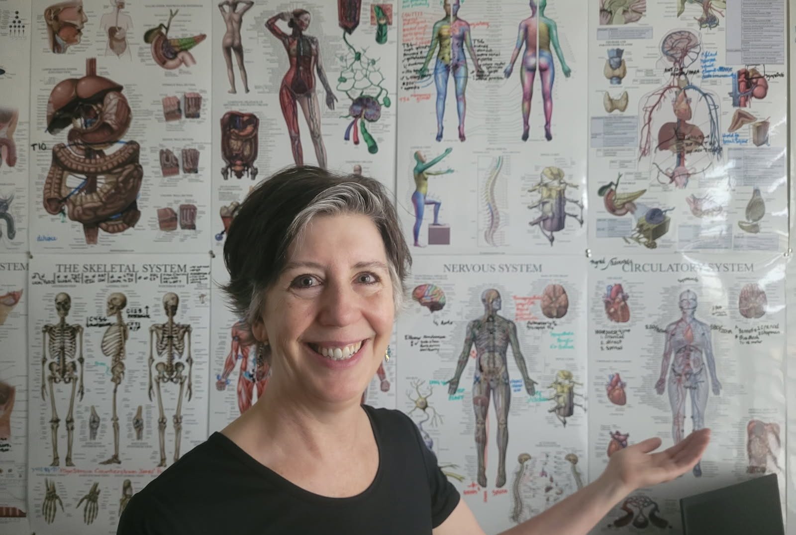 Rachel Tobler smiling in front of a wall of anatomy posters showing the skeletal, nervous and circulatory systems