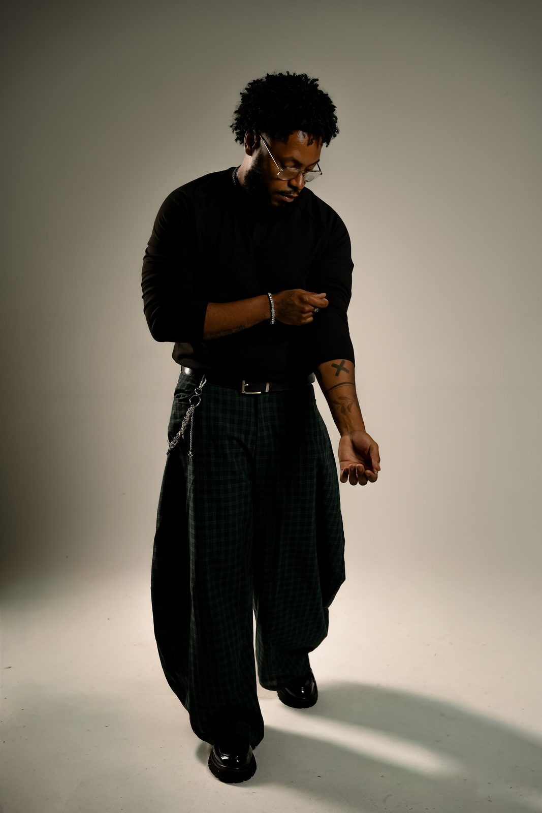 Remy Janvier standing in a photo studio in a black top and green plaid trousers, looking down at his open hand
