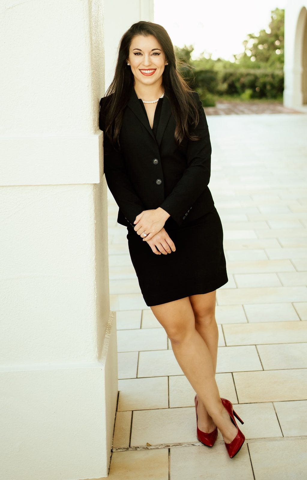 Sara Varnado in a black suit and red heels, leaning against a white column on a sunlit terrace