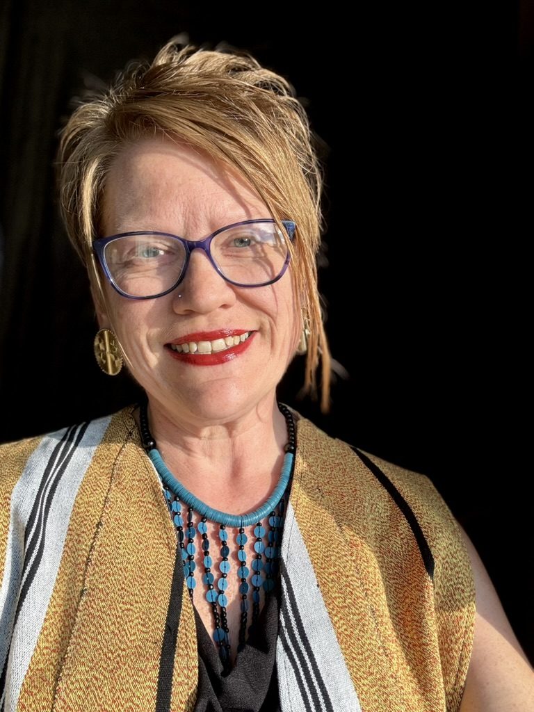 Sarah Drake, founder of herARTS in Action, smiling in purple glasses, red lipstick and a mustard striped shawl with a turquoise beaded necklace against a dark background