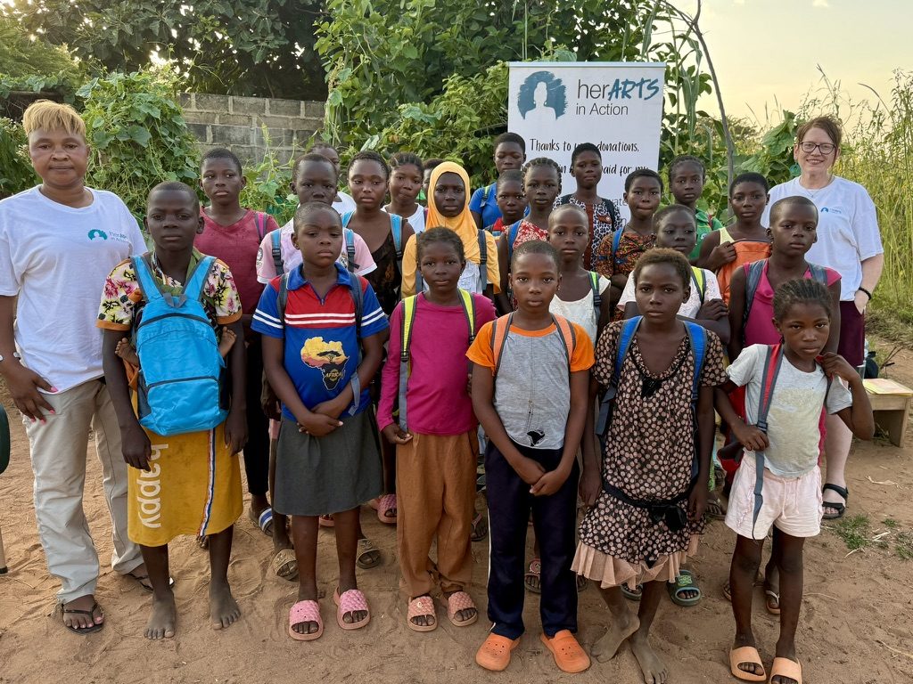 Sarah Drake, right, in a white herARTS in Action shirt with a large group of girls carrying backpacks in front of a herARTS in Action banner in Burkina Faso