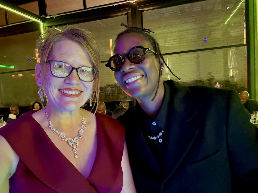Sarah Drake in a burgundy gown and crystal necklace smiling beside a guest in a black suit and round glasses at an evening gala