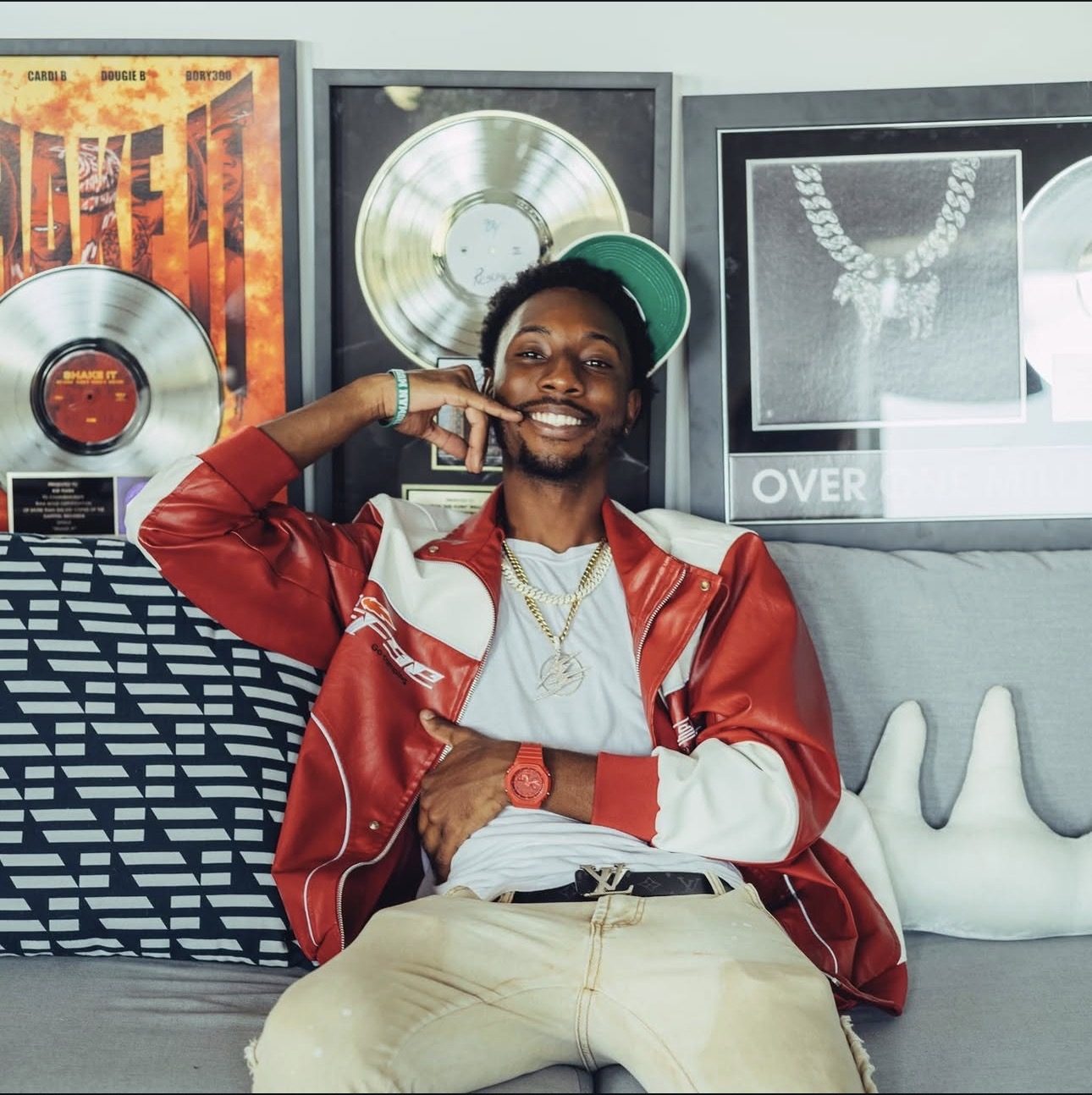 Sean "Kid Flvsh" Williams, founder of Flash FWD Productions, smiling on a sofa in a red and white jacket in front of framed platinum records
