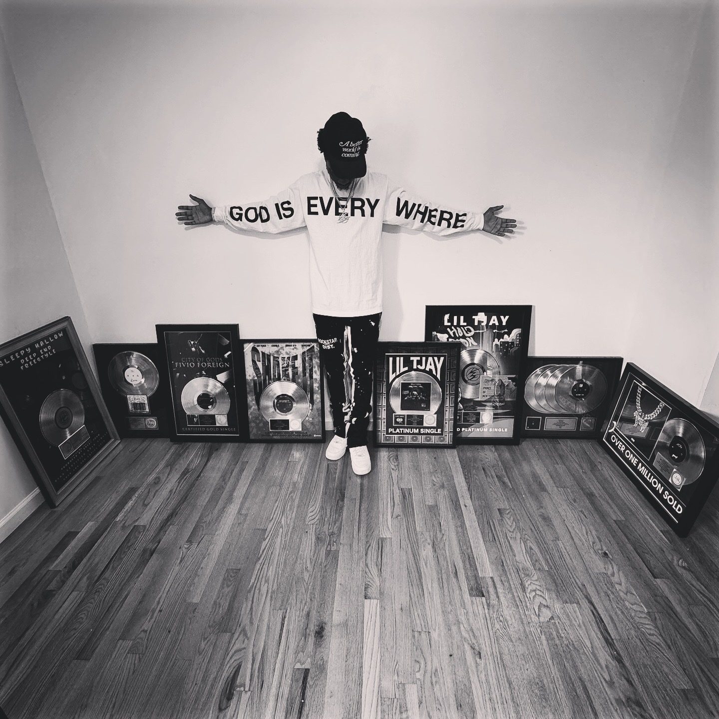 Black and white photo of Kid Flvsh with arms outstretched in a God Is Everywhere shirt, surrounded by framed platinum and gold plaques