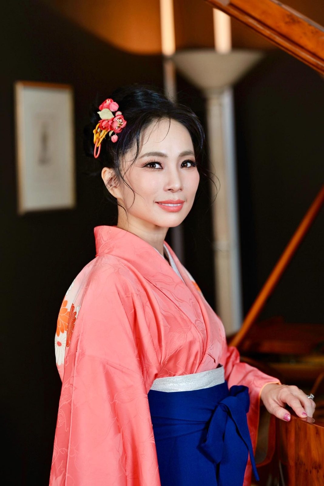Shiho Brenner in a coral kimono and navy hakama with flowers in her hair, standing beside a grand piano