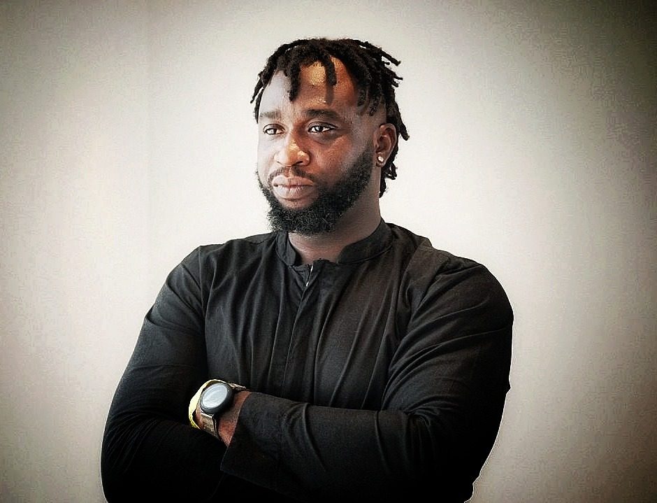 Simpa Samson, documentary filmmaker and founder of VisualFarm Production, arms crossed in a black shirt against a pale wall