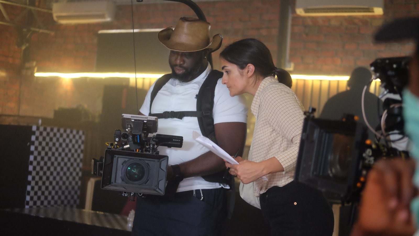 Simpa Samson in a brown hat holding a cinema camera beside a colleague reading a script on a film set