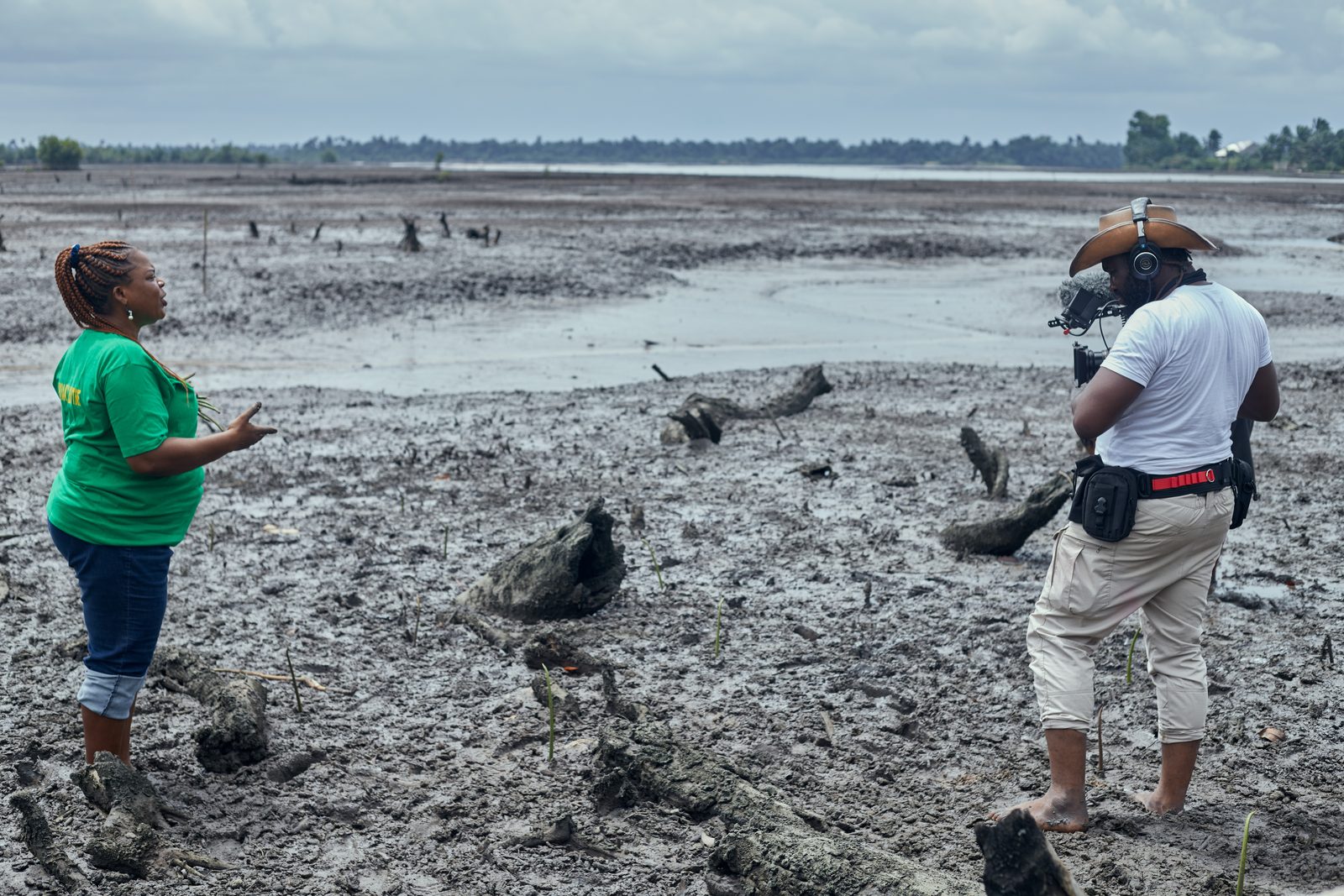 Simpa Samson filming a woman in a green T-shirt as she speaks on a mudflat of felled tree stumps
