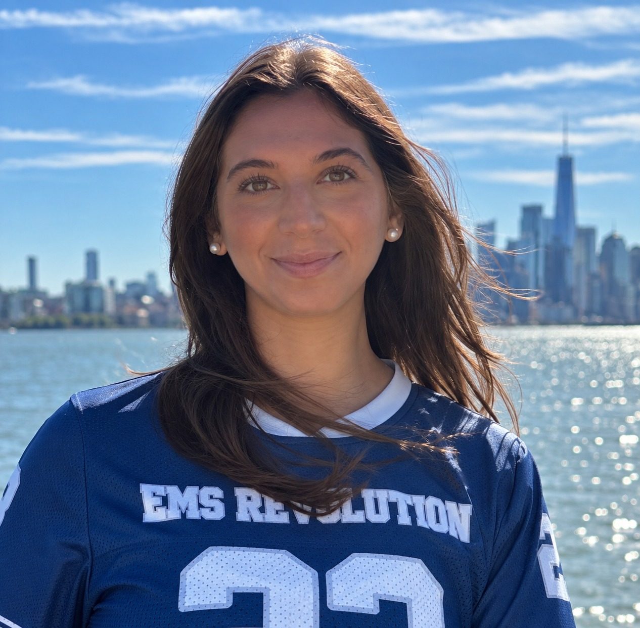 Sydney Kessler, founder of EMS Revolution, in a navy EMS Revolution jersey on the Hudson waterfront with the Manhattan skyline behind her