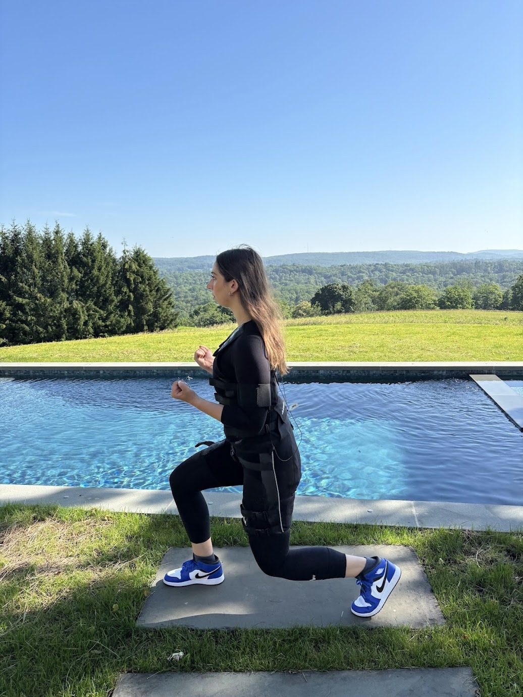 Sydney Kessler in an EMS training suit doing a lunge beside a pool, with wooded hills in the distance