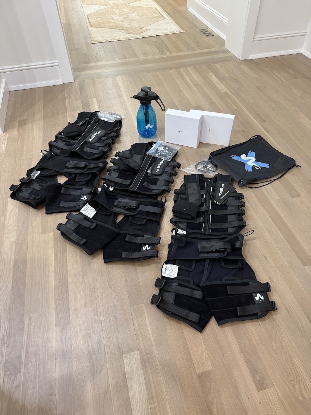 EMS training vests, shorts and arm straps laid out on a hardwood floor with a spray bottle, boxed equipment and a drawstring bag