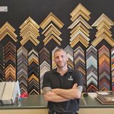 Adam Fins, owner of Friedman's Framing in Savannah, Georgia, standing with arms crossed at the shop counter in front of a wall of frame corner samples