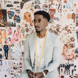 Adeola Odutola, founder and creative director of Crown of Wealth, seated in a pale suit with a tape measure around his neck in front of a wall of his designs