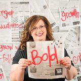 April Barnard, founder of Marketing with April, smiling and holding up a newspaper page marked "Bold" in red, in front of a wall of newspapers