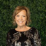Goldie Benz, owner of GoldenWorks Embroidery, smiling in a black top with silver sequined leaves against a green hedge wall