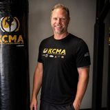 Keith Kirkpatrick, founder of Kombative Concepts Martial Arts, smiling beside a heavy bag in a KCMA T-shirt