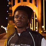 Keysean Jenkins, founder of Dios Primero, in a black Dios Primero jacket reading Halleluyah Praise Yah on a lit bridge in Pittsburgh at night