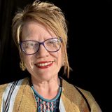 Sarah Drake, founder of herARTS in Action, smiling in purple glasses, red lipstick and a mustard striped shawl with a turquoise beaded necklace against a dark background