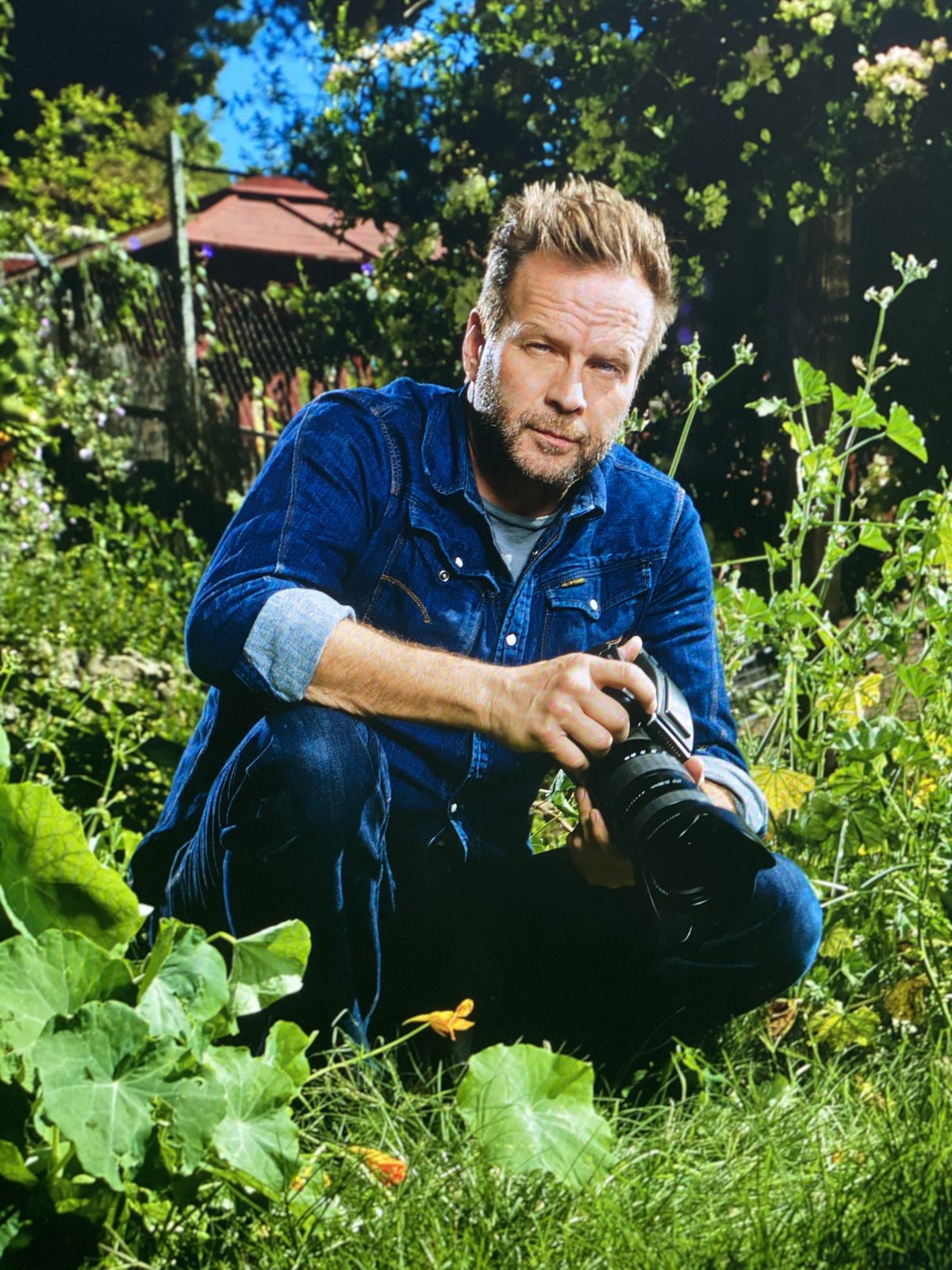 William Sanford, founder of H.I.P Productions, kneeling in a Los Angeles garden in a denim shirt with his camera in hand